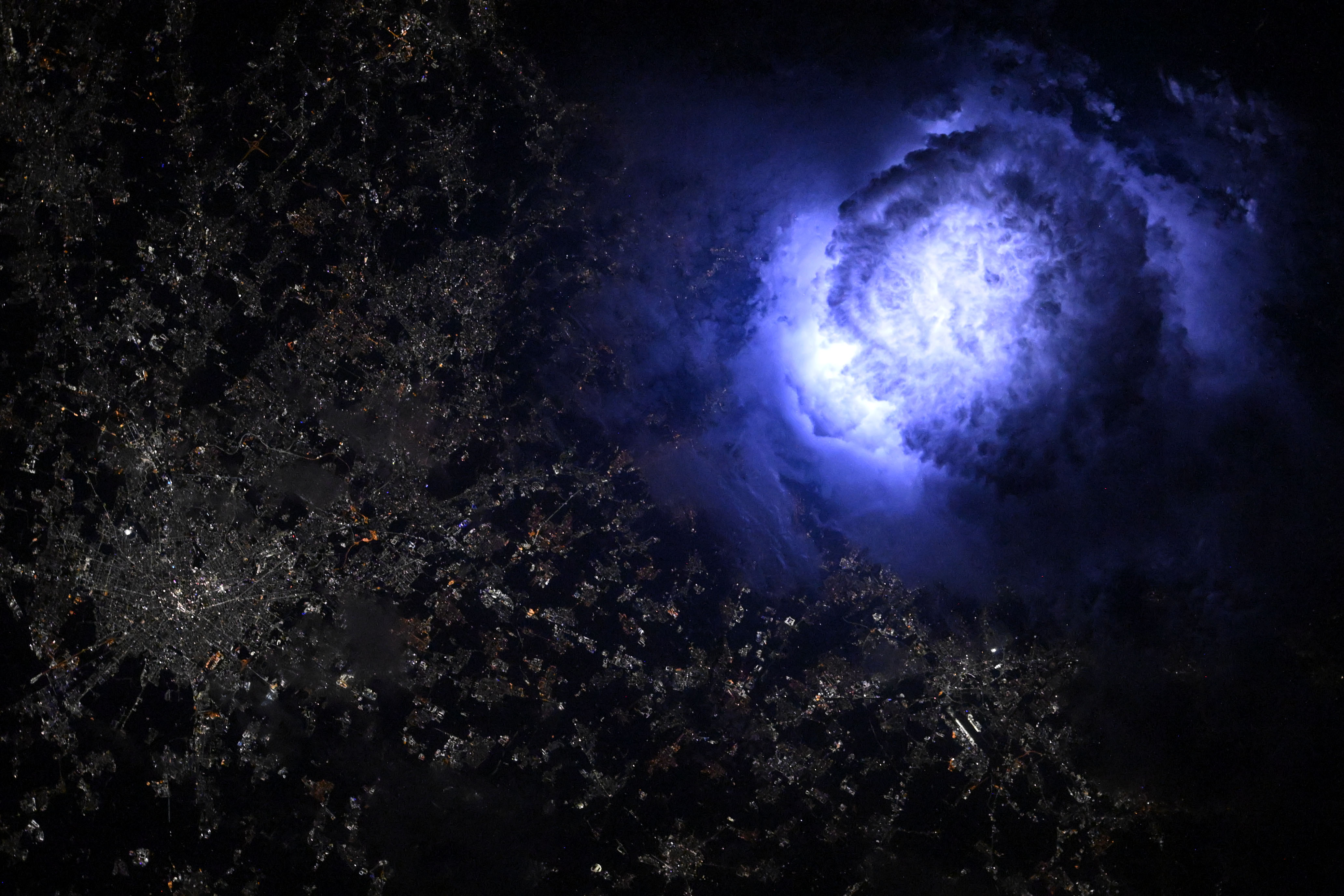 An enormous cloud glows blue-white with lightning over a web of city lights in northern Italy.